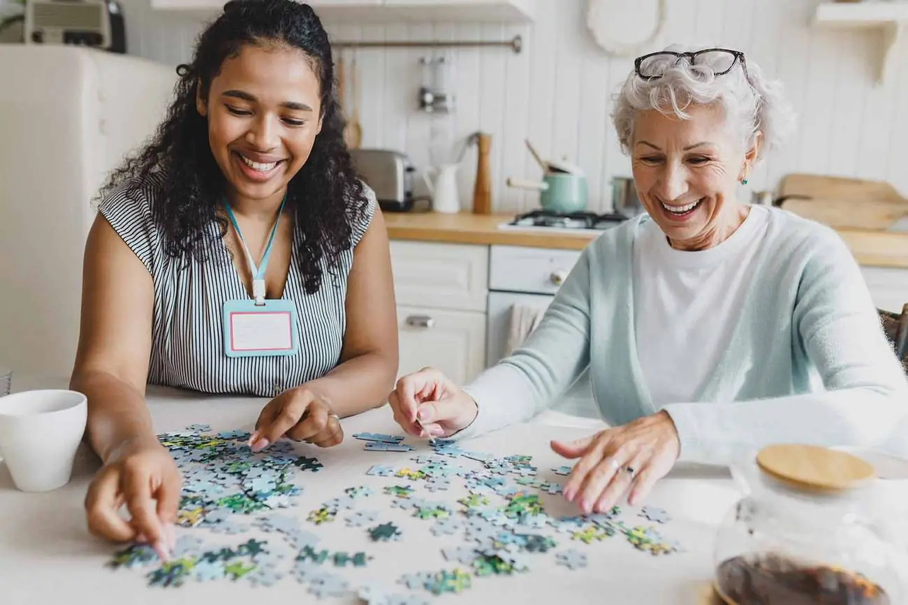 A carer with a lanyard and an older woman doing a jigsaw together at a kitchen table, both smiling